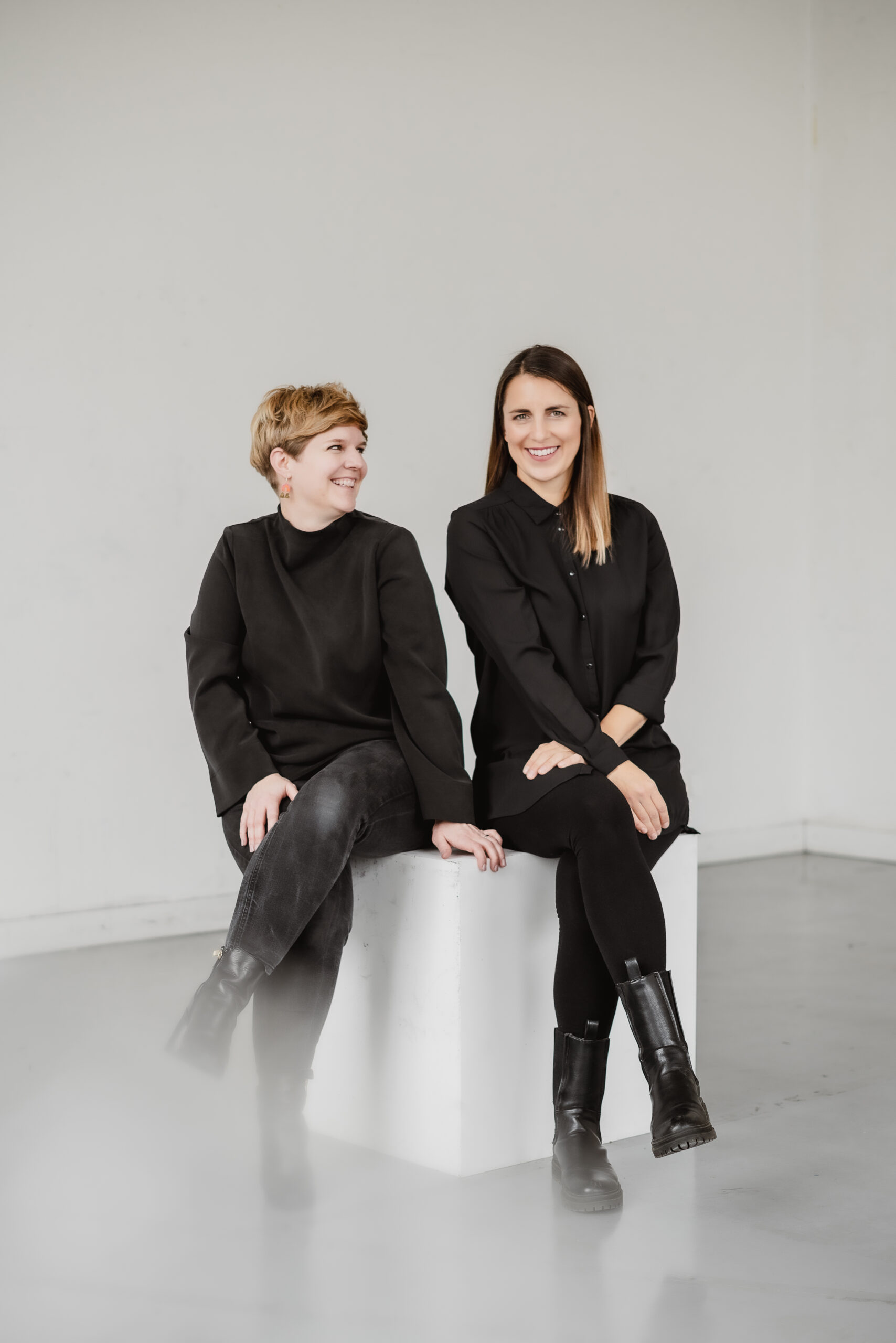 Die Geschäftsführerinnen Isabelle Possehl und Janina Schneider sitzen vor einer weißen Wand nebeneinander auf einem weißen Würfel. Isabelle Possehl schaut lächelnd zu Janina Schneider rüber, welche auch lächelnd in die Kamera schaut.